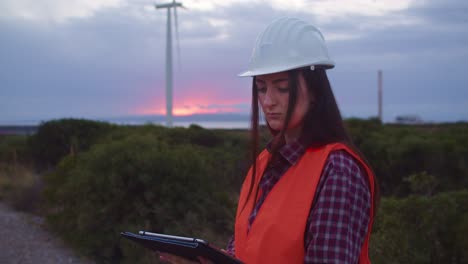 Woman-engineer-checks-tablet-to-inspect-wind-turbines-parameters,-slomo,-sunset