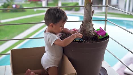 Junger-Lateinischer-Junge,-Der-Auf-Dem-Dreck-In-Einem-Blumentopf-Spielt,-Mit-Seinem-Autospielzeug,-Das-In-Einem-Karton-Sitzt