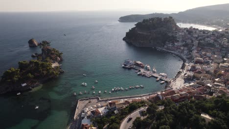 Aerial-view-of-coastal-Greek-town-Parga,-ships-anchored-in-city-dock,-people-walk-along-promenade,-orbiting