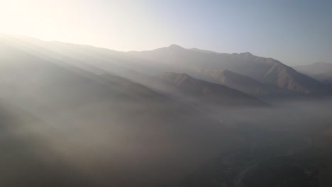 Eine-Seitliche-Drohnenaufnahme-Von-Nebligen-Bergen-Bei-Sonnenuntergang-Mit-Riesigen-Lichtstrahlen-In-Peru