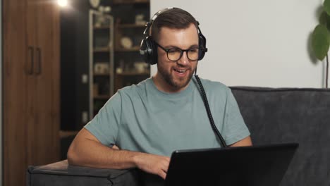 Handsome-man-in-glasses-puts-on-headphones-and-waves-hello-to-laptop-webcam