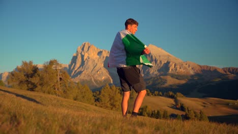 Mountains,-forest-and-grass-fields-with-a-man-holding-an-Italian-flag-filmed-at-Alpe-di-Siusi-in-Alps,-Italian-Dolomites-filmed-at-sunset