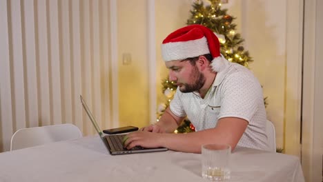 Junger-Kaukasischer-Mann-Mit-Weihnachtsmütze,-Der-Auf-Einem-Laptop-In-Seinem-Wohnzimmer-Mit-Einem-Weihnachtsbaumglühen-Im-Hintergrund-Konzentriert-Schreibt