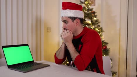 Frightened-young-man-with-Santa-hat-sitting-at-table-next-to-Christmas-tree-and-looking-at-a-laptop-green-screen