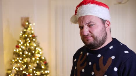 Frowning-young-man-with-Santa-hat-and-a-Xmas-sweater-standing-in-front-of-a-Christmas-tree-with-a-disappointed-expression-on-his-face