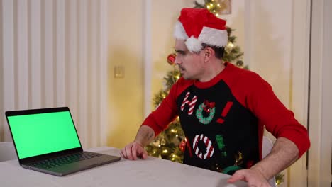 Hombre-Feliz-Con-Sombrero-De-Santa-Se-Sienta-A-Hablar-Con-La-Familia-Sobre-Zoom-Skype-Durante-Navidad,-Mira-Un-Video-En-Una-Pantalla-Verde-De-Computadora-Portátil