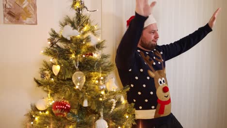 Young-happy-man-with-a-Rudolf-Xmas-jumper-and-a-Santa-hat-dancing-to-the-music-while-standing-near-glowing-Christmas-tree