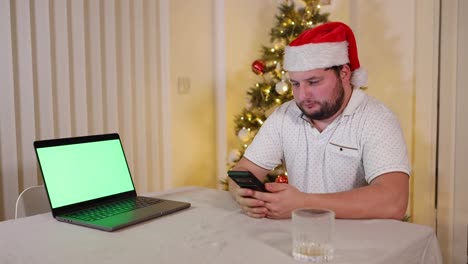 Concentrated-focused-man-sitting-on-a-table-scrolls-his-phone,-looks-to-camera-and-to-laptop-with-green-screen,-wearing-Santa-hat