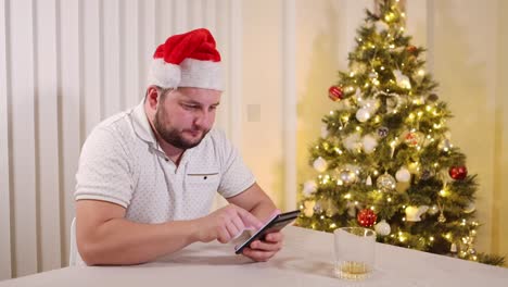 Impressed-and-happy-man-sitting-near-a-decorated-Christmas-tree-and-scrolling-on-a-smartphone,-happy-smile