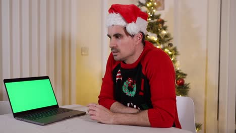 Hombre-Con-Un-Sombrero-De-Santa-Sentado-Cerca-De-Una-Computadora-Portátil-Con-Pantalla-Verde-Y-Pensando,-Tiempo-De-Navidad