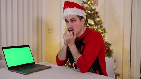 Man-with-Christmas-clothes-sitting-next-to-xmas-decorated-tree-looking-shocked-to-laptop-screen-with-green-screen-chroma-key