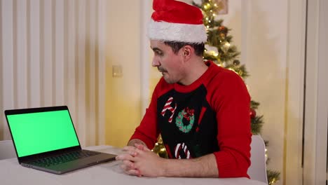 Un-Hombre-Caliente-En-Un-Saltador-De-Navidad-No-Puede-Creer-Lo-Que-Ha-Visto-En-La-Pantalla-Verde-De-Una-Computadora-Portátil,-árbol-De-Navidad-Decorado-En-El-Fondo