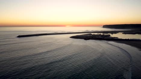 Schließende-Drohnenaufnahme-Von-Wellen,-Die-Während-Des-Sonnenuntergangs-Auf-Einen-Strand-Mit-Einem-Fischerhafen-Im-Hintergrund-Stürzen