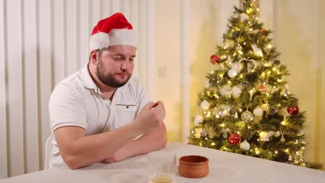 Young-man-sitting-next-to-a-decorated-xmas-tree-eating-Christmas-snack-while-wearing-Santa-hat,-static-view