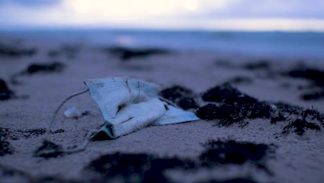 Used-face-mask,-trash-and-waste-litter-on-an-empty-Baltic-sea-white-sand-beach,-environmental-pollution-problem,-overcast-evening-after-sunset,-closeup-shot