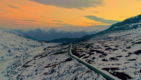 Aerial-pullback-over-road-cutting-through-snowy-mountainous-landscape---vibrant-orange-sunset-sky-in-Norway