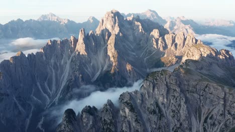 Aerial-wide-shot-of-sunlight-bathing-a-mountain-range-in-Dolomites-in-the-morning---Dense-Clouds-covering-valley