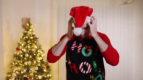 Man-wearing-festive-jumper-in-front-of-Christmas-tree-thrown-Santa-hat-and-blows-snowball-end-off-his-face-pointing-at-camera-and-smiling