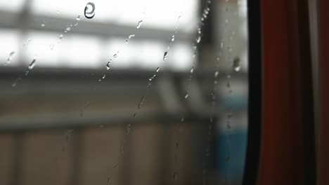 Water-dripping-down-on-the-window-glass,-blurry-train-station-background,-closeup