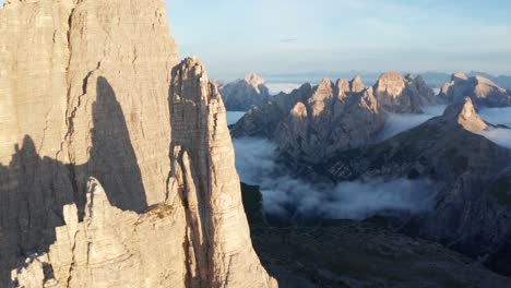 Cinematic-drone-shot-showing-Tre-Cime-spires-in-Dolomite-Mountains-during-a-golden-sunrise---Clouds-covering-valley-in-background---Panoramic-shot