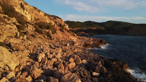 Luftaufnahme-Küstenfelsen-Der-Bucht-Von-Cala-Xarraca-Auf-Ibiza,-Spanien,-Während-Die-Wellen-Auf-Den-Felsen-Mit-Bergen-Und-Blauem-Himmel-Im-Hintergrund-Krachen