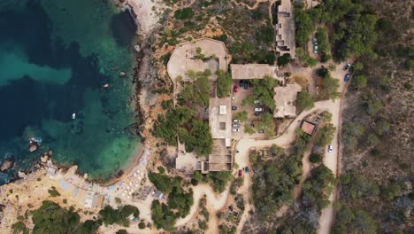 Aerial-top-view-of-abandoned-hotel-Sert-at-Cala-d’en-Serra-Beach,-Spain-and-clear-blue-ocean-with-rock-cliffs,-roads-and-trees