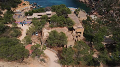 Abandoned-hotel-on-cliff-top-at-Cala-d’en-Serra-Beach,-Spain-with-graffiti,-trees,-roads-and-ocean-cove-in-background---Drone-aerial-flyover-footage