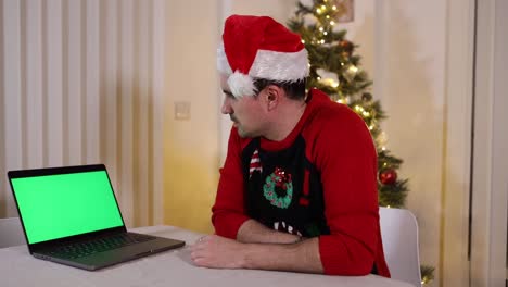 casually-dressed-businessman-impatiently-looking-at-the-green-screen-of-his-laptop-in-home-office-during-the-Christmas-holiday-wearing-a-Santa-hat