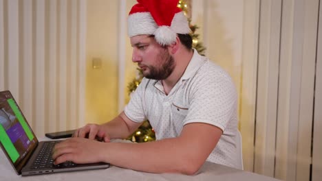 Fokussierter-Geschäftsmann-Unternehmer-Mit-Weihnachtsmütze,-Der-Während-Der-Weihnachtsferien-In-Seinem-Heimbüro-Mit-Geschmücktem-Weihnachtsbaum-Im-Hintergrund-An-Einem-Laptop-Arbeitet