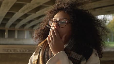 Schwarze-Frau,-Die-Während-Ihres-Herbstspaziergangs-Draußen-Unter-Einer-Brücke-Eine-Zigarette-Raucht