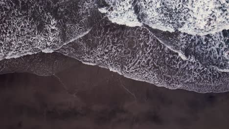 Aerial-top-down-view-of-wave-rolling-on-black-sand-beach,-background