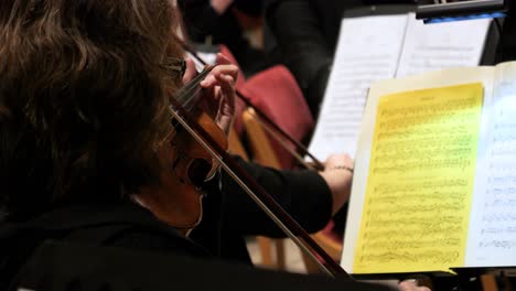 Close-up-of-a-female-violin-player-performing-with-a-wooden-violin-with-orchestra-during-a-concert-with-classical-music-to-mozart-requiem-with-view-of-other-violin-players-and-sheet-music