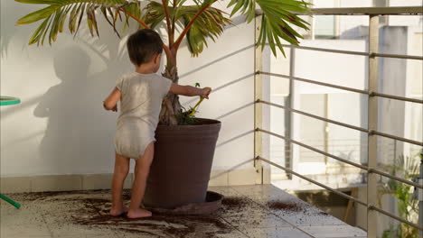 Little-latin-baby-boy-scooping-dirt-from-a-plant-pot-and-dropping-it-to-the-ground-with-a-green-plastic-kitchen-spoon