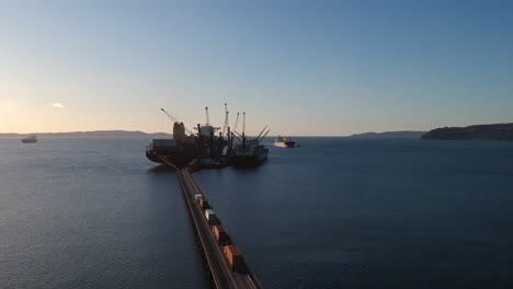 Trucks-are-moving-over-the-bridge-into-industrial-shipping-port,-aerial-view