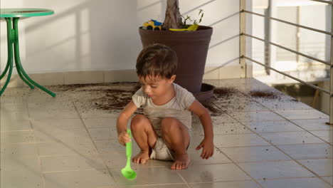 Freches-Kleines-Kleinkind,-Das-Mit-Einem-Grünen-Plastiklöffel-Spielt,-Nachdem-Es-Den-Ganzen-Boden-Mit-Schmutz-Aus-Einem-Blumentopf-Verschmutzt-Hat