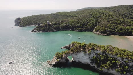 Drone-flying-backwards-with-the-camera-tilting-up-slowly-showing-an-arch-in-the-sea-at-the-coast-of-Gargano-national-park,-Italy-in-4k