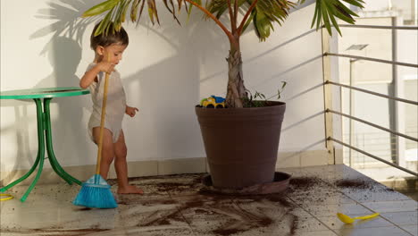 Little-latin-baby-boy-wearing-a-baby-romper-cleaning-the-floor-with-a-blue-toy-broom-after-soiling-it-with-dirt-from-a-plant-pot