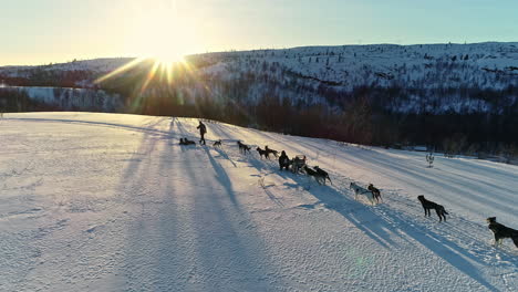 People-and-sled-dogs-stopped-on-snow-with-sunbeams-behind-mountains-at-sunset