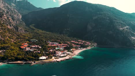 Luftaufnahme-Eines-Resorts-Am-Strand-Mit-Bergen-Im-Hintergrund-Und-Einem-Vorbeifahrenden-Boot---Ölüdeniz---Türkei