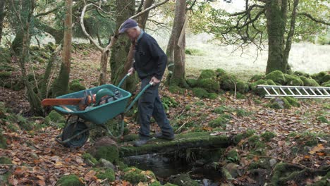 Mature-man-crosses-a-stream-with-a-wheelbarrow-loaded-with-a-chainsaw