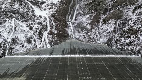 Amazing-drone-footage-over-an-alpine-lake,-hydro-dam-and-vast-Alpine-reservoir-fringed-by-rugged-mountains-with-a-waterside-trail-after-the-first-snow-fall-of-winter-2022