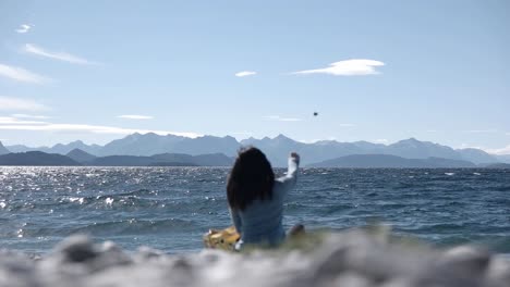 Woman-sitting-On-Shoreline-And-Throwing-Stones-In-Nahuel-Huapi-Lake-On-Clear-Sunny-Day