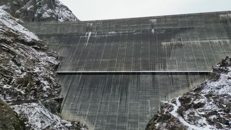 Amazing-drone-footage-over-an-alpine-lake,-hydro-dam-and-vast-Alpine-reservoir-fringed-by-rugged-mountains-with-a-waterside-trail-after-a-light-snow-fall