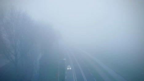 Ein-Luftbild-Einer-Mit-Mystischem-Nebel-Bedeckten-Straße-Im-Dunkeln-Und-Vorbeifahrenden-Autos