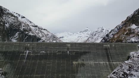 Amazing-drone-footage-over-an-alpine-lake,-hydro-dam-and-vast-Alpine-reservoir-fringed-by-rugged-mountains-with-a-waterside-trail-after-a-light-snow-fall