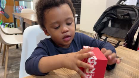 Frustrated-and-funny-black-two-year-old-baby-tries-to-open-a-tiny-red-gift-box-seated-next-to-mum-inside-a-cafeteria