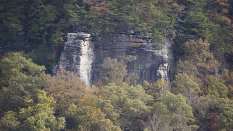 Static-shot-of-a-large-cliff-face-surrounded-by-autumnal-trees-in-the-mountains
