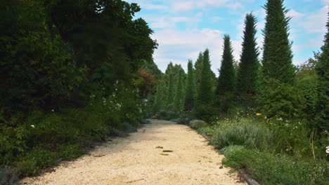 centered-orange-gravel-european-pathway-turns-the-curve,-left-and-right-along-the-track-surrounded-by-mediterranean-green-shrubs-and-plants,-blue-clean-sky-above-filled-with-some-summer-clouds
