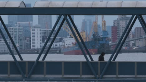 Man-Walking-Over-Bridge-with-River-Thames-Industry,-Cargo-Ship-and-Financial-District-in-Background