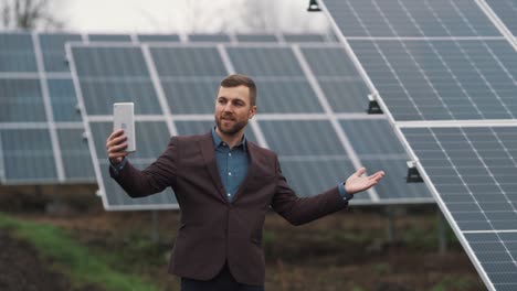 A-solid-man-in-business-clothes-is-talking-with-a-tablet-on-the-background-of-a-power-plant-made-of-solar-panels